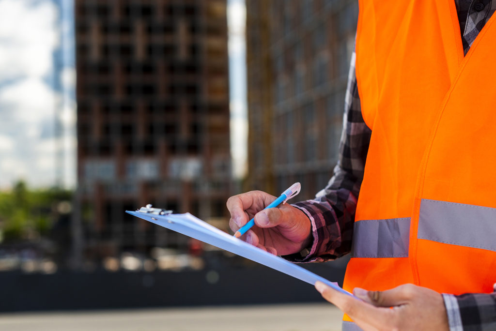 Construction contractor in an orange safety vest reviewing a clipboard on a job site, with a partially constructed building in the background, emphasizing the assessment phase of property reconstruction services.